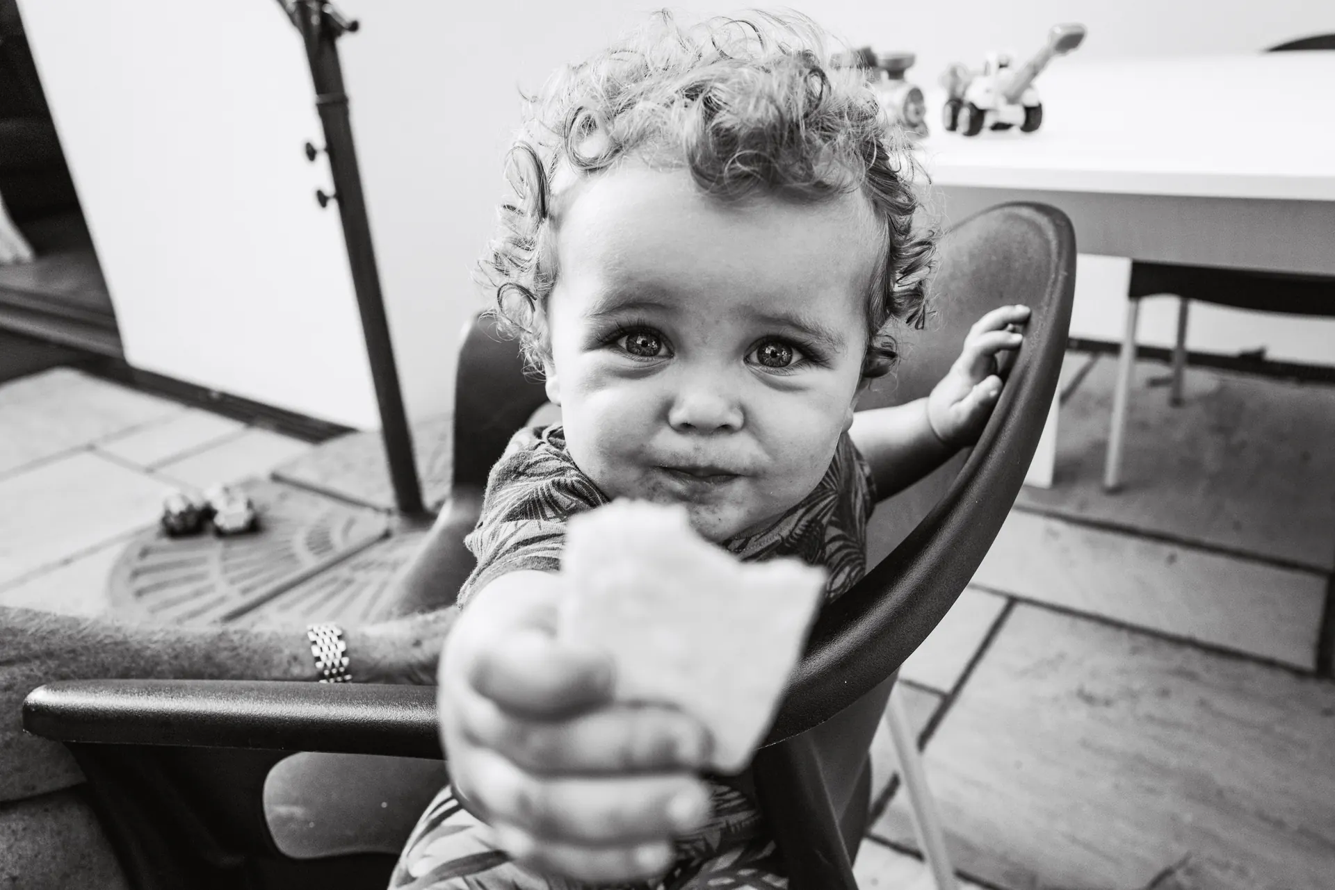 Toddler in a high chair offering a biscuit to the camera