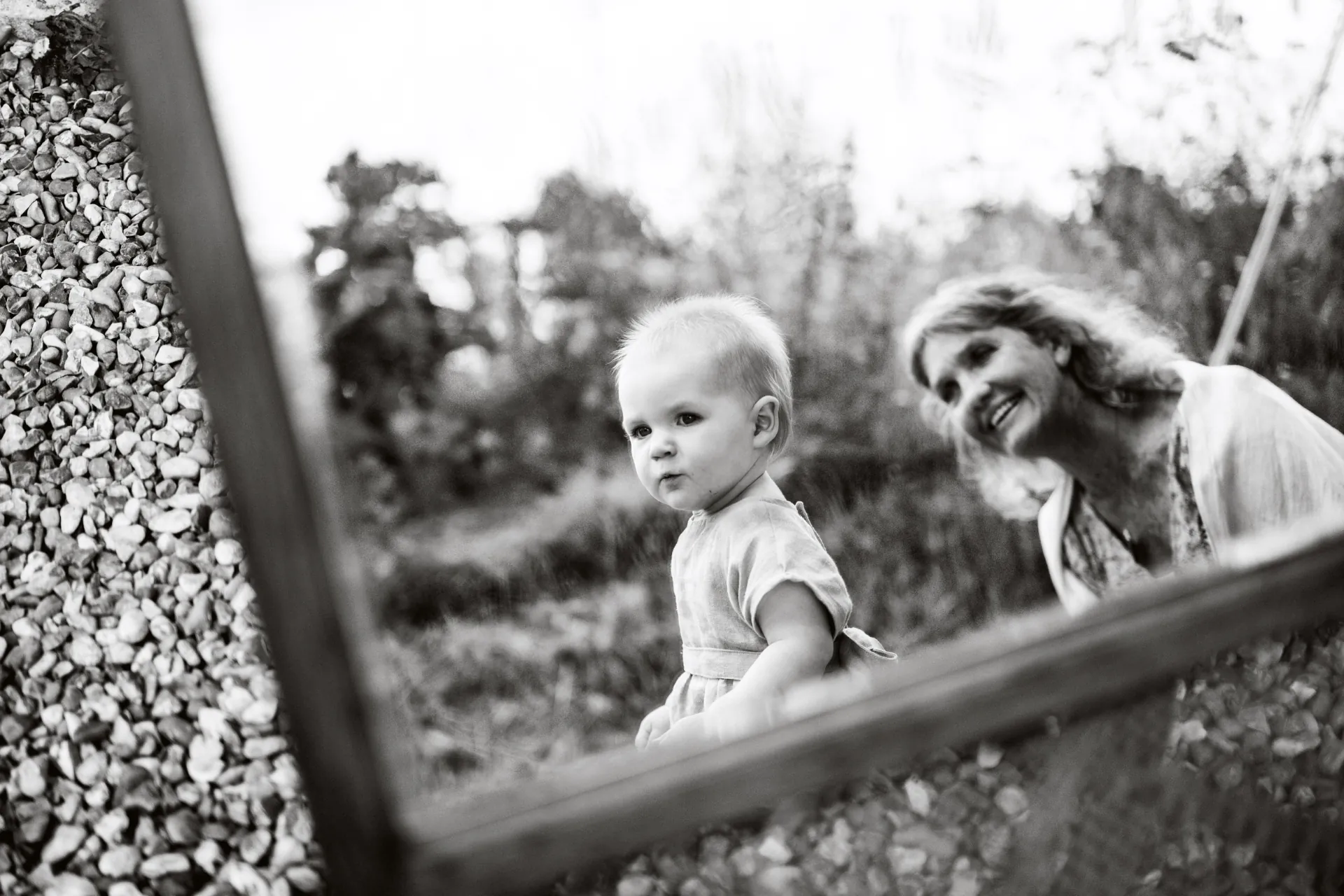 Black and white photo of a toddler in a garden with a smiling grandparent behind