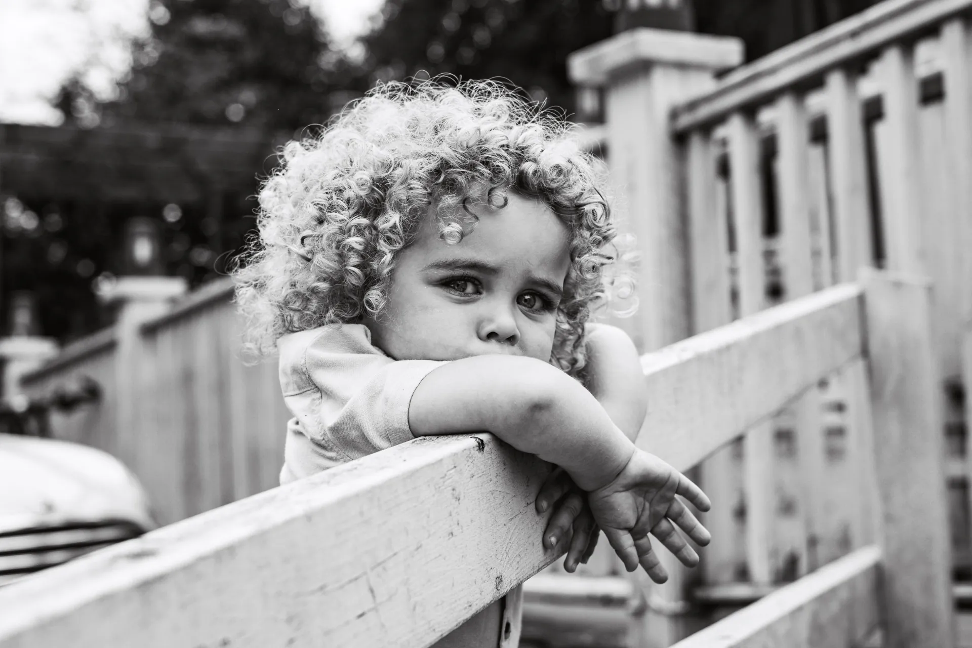 Black and white portrait of a curly-haired child leaning on a garden fence outdoors