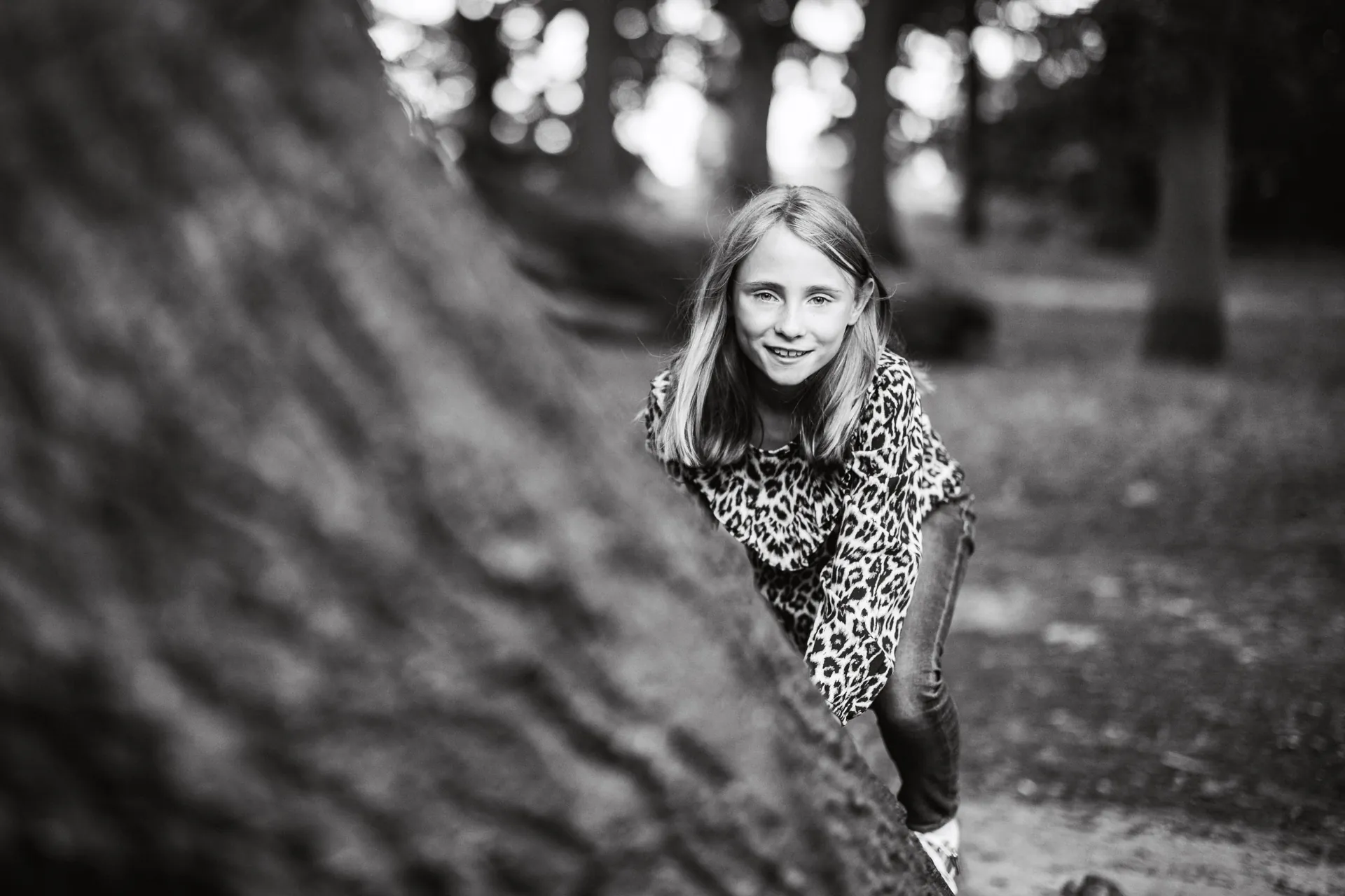 Black and white portrait of a smiling girl climbing over a fallen tree in woodland