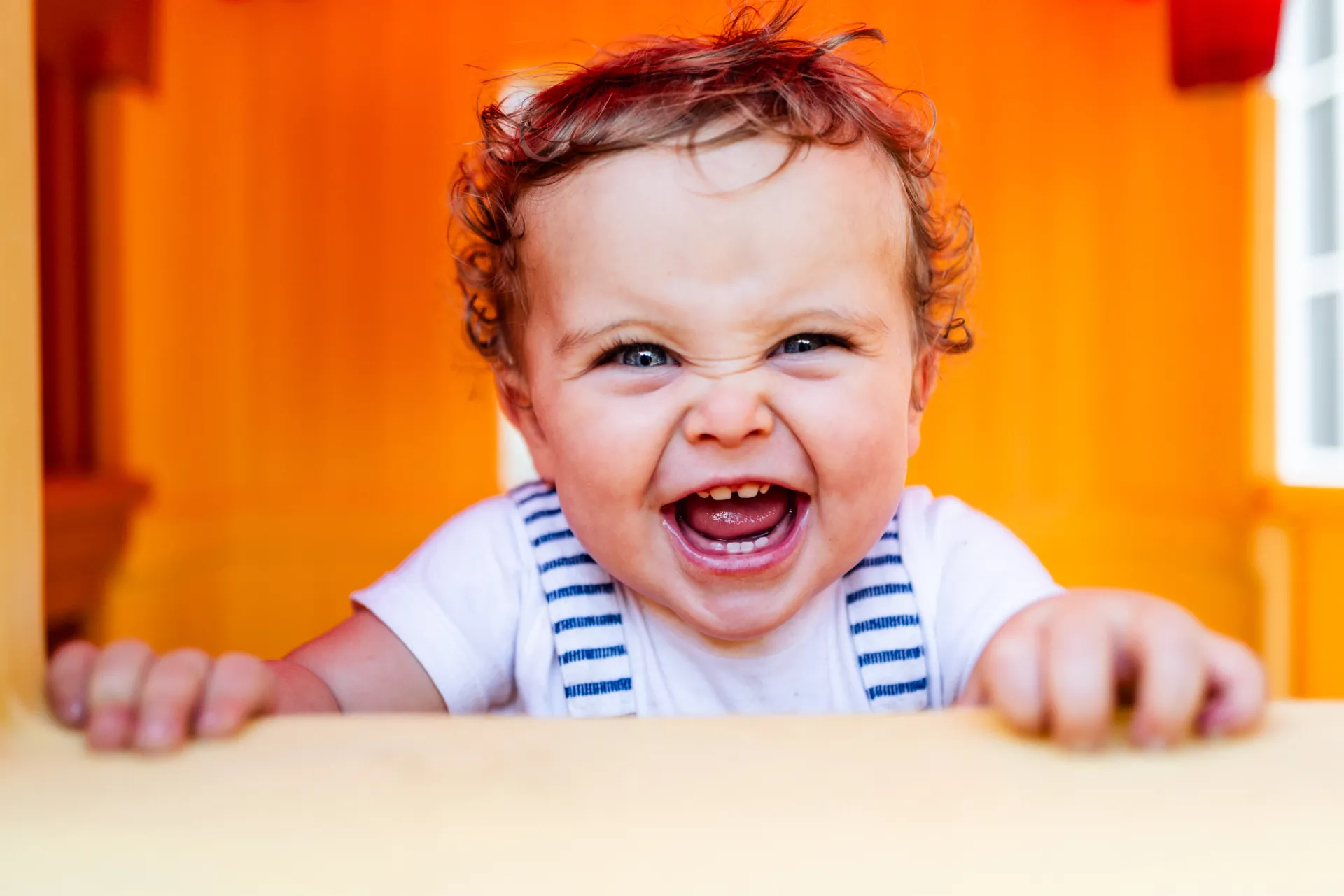 A laughing toddler peeking over the edge of a bright orange playhouse