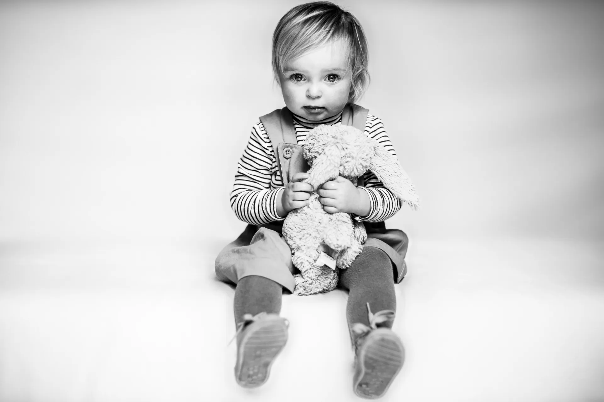 Black and white studio portrait of a toddler sitting on the floor hugging a soft toy